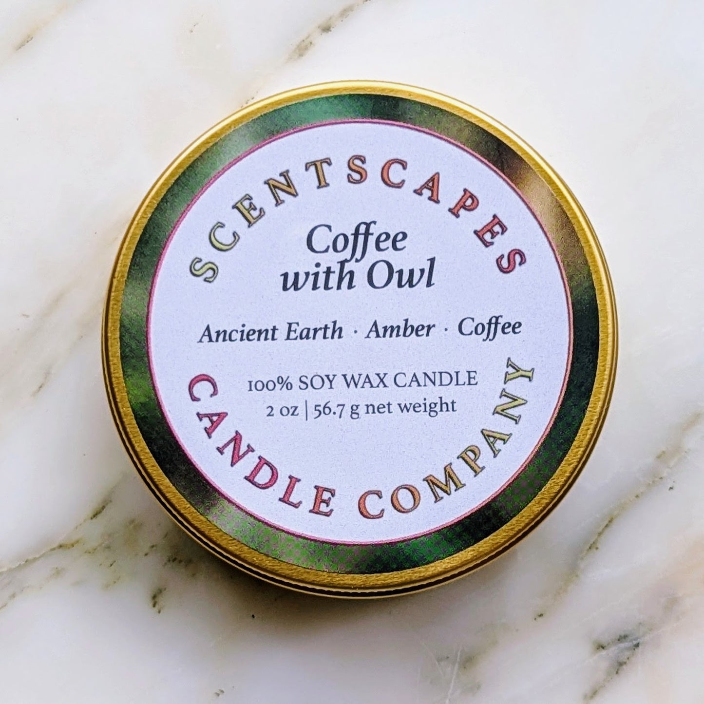 Scentscapes small candle in gold tin labeled 'Coffee with Owl' with scents Ancient Earth, Amber, Coffee on a marble surface.