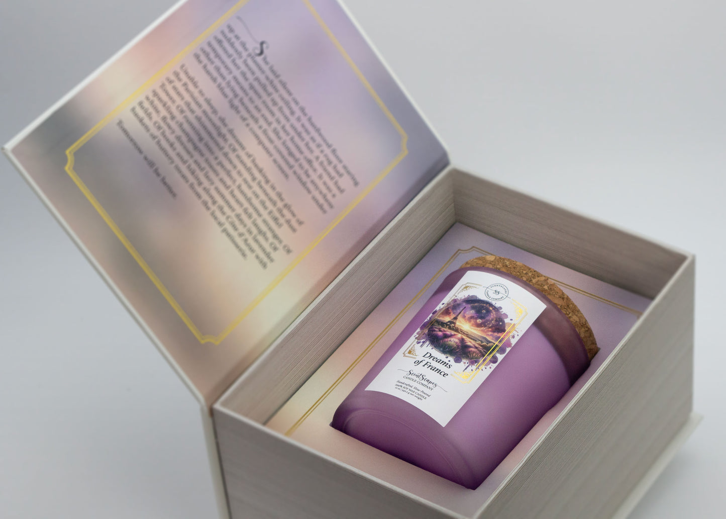A ScentScapes candle in a purple jar with a cork lid, labeled 'Dreams of France' with an image of a glittering the Eiffel Tower and lavender fields. Candle inside its storybook box packagine. Unboxing image reveals short story inside front cover that goes along with the scent.