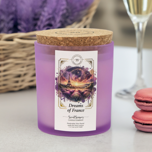 A ScentScapes candle in a purple jar with a cork lid, labeled 'Dreams of France' with an image of a glittering the Eiffel Tower and lavender fields. A vintage-style gold foil design frames the image. The candle is displayed with a basket, glass of champagne and macarons in the background.