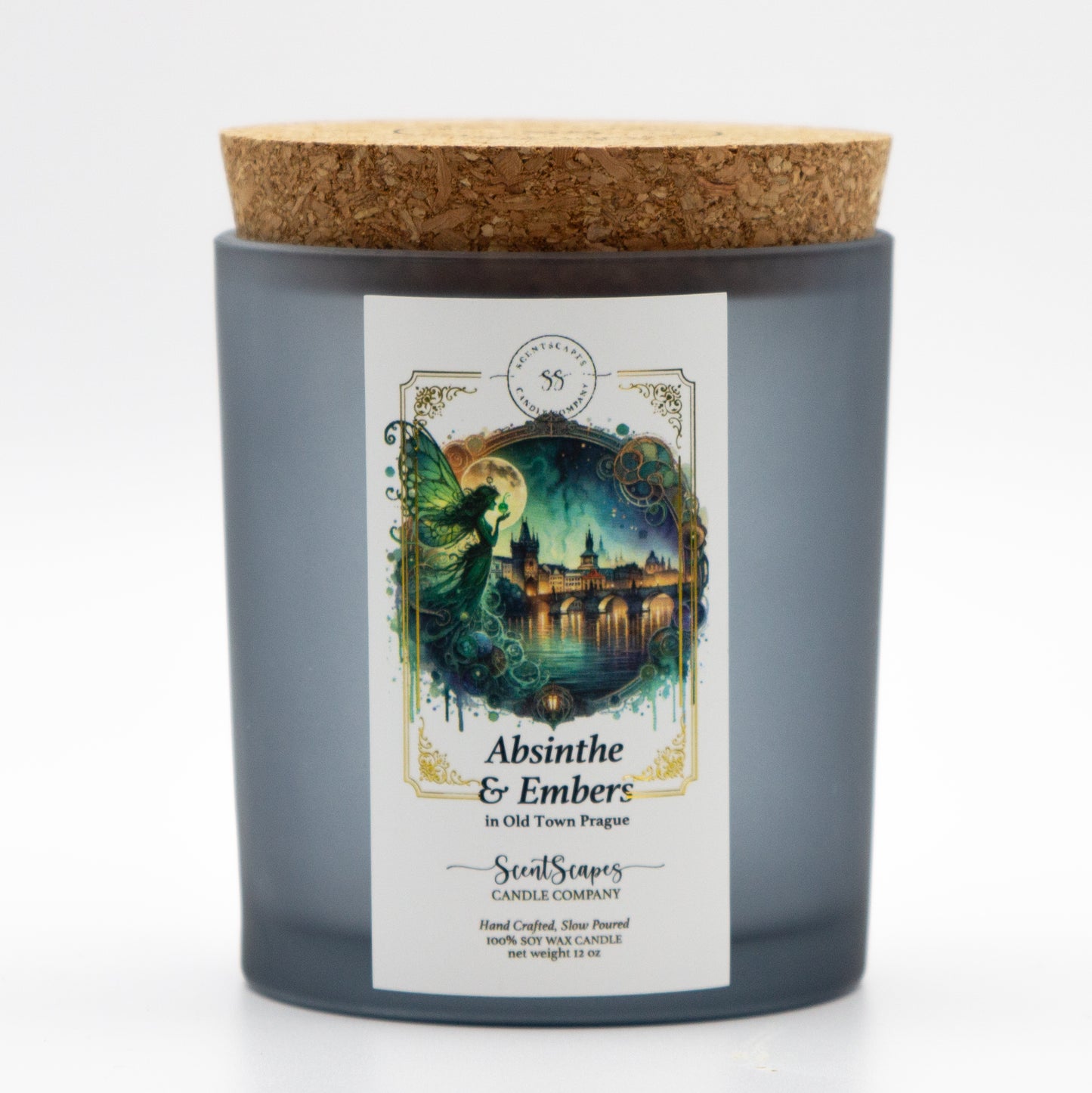 A lit ScentScapes candle in a gray black glass jar and cork lid with a label featuring an illustration of a green fairy framed by a full moon with Old Town Prague in the background and Charles Bridge along the Vltava River. A vintage-style gold foil design frames the image.
