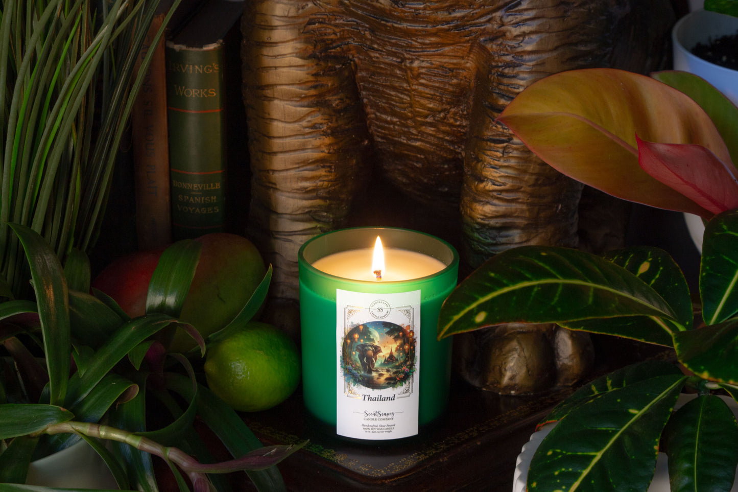 A lit ScentScapes scented candle in green jar with a cork lid, labeled 'Thailand', depicting an illustration of a sunset jungle scene with lush foliage and glowing lanterns. Features an elephant walking through a stream of water with an ancient Thai temple in the background.