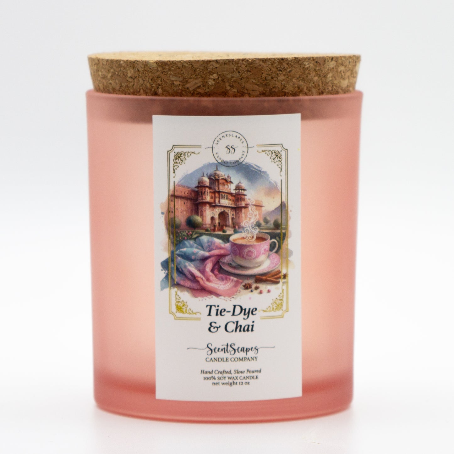 A ScentScapes candle in a pink glass jar with a cork lid, featuring an illustration of a cup of chai, pastel pink and blue tie-dye fabric and the pink city of Jaipur, India in the background. A vintage-style gold foil design frames the image.