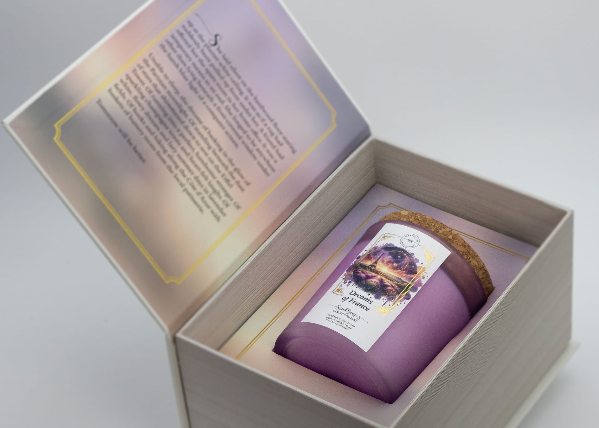 A ScentScapes candle in a purple jar with a cork lid, labeled 'Dreams of France' with an image of a glittering the Eiffel Tower and lavender fields. Candle inside its storybook box packagine. Unboxing image reveals short story inside front cover that goes along with the scent.