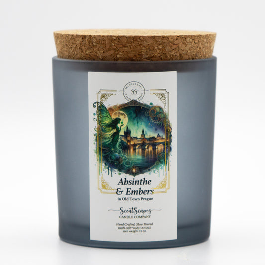 A lit ScentScapes candle in a gray black glass jar and cork lid with a label featuring an illustration of a green fairy framed by a full moon with Old Town Prague in the background and Charles Bridge along the Vltava River. A vintage-style gold foil design frames the image. 