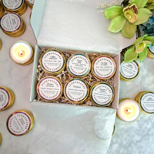 Box of Scentscapes candles in gold sample tins as part of their Scent Discovery Kit featuring a variety of scents. On top of white marble surface surrounded by lit candles.
