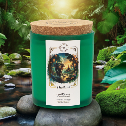 A ScentScapes scented candle in green jar with a cork lid, labeled 'Thailand', depicting an illustration of a sunset jungle scene with lush foliage and glowing lanterns. Features an elephant walking through a stream of water with an ancient Thai temple in the background. A vintage-style gold foil design frames the image.