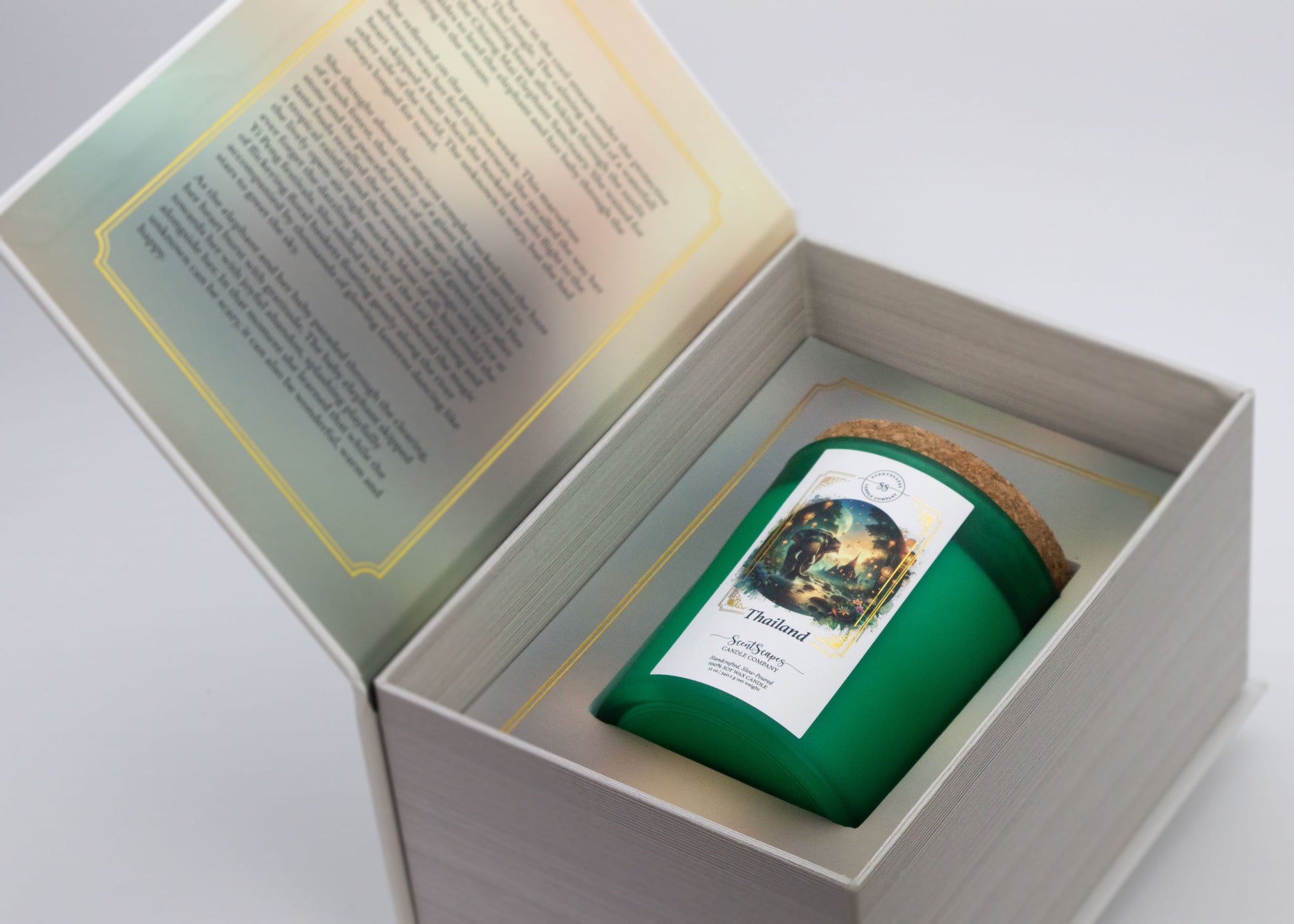 A ScentScapes scented candle in green jar with a cork lid, labeled 'Thailand', depicting an illustration of a sunset jungle scene with lush foliage and glowing lanterns. Features an elephant walking through a stream of water with an ancient Thai temple in the background. The candle is inside its storybook box packaging. The unboxing image reveals a short story on the inside cover that goes along with the scent.